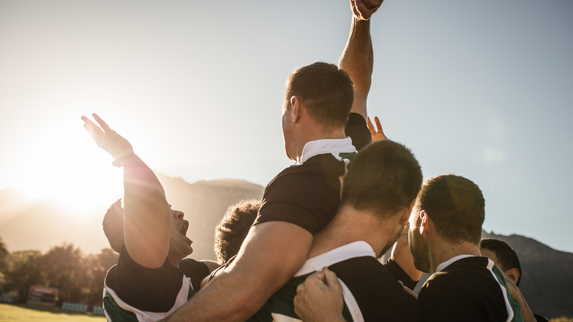 A group of male rugby players celebrate on the field, raising each other up after a game.